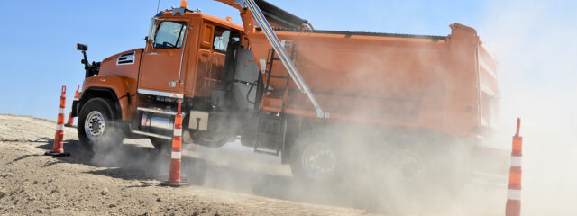 An orange dump truck with a lift axle driving up a dirt incline