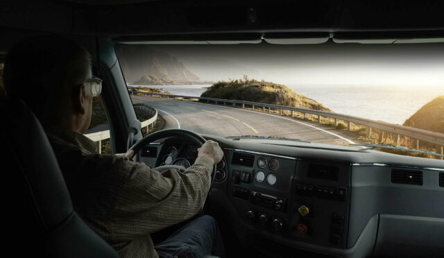 A photo taken from behind a driver in a semi cab looking out the windshield at a road and a view of the ocean