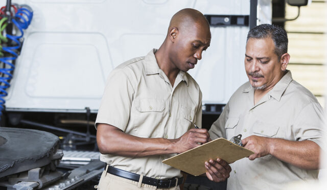 Two men looking at a clipboard behind a white semi truck