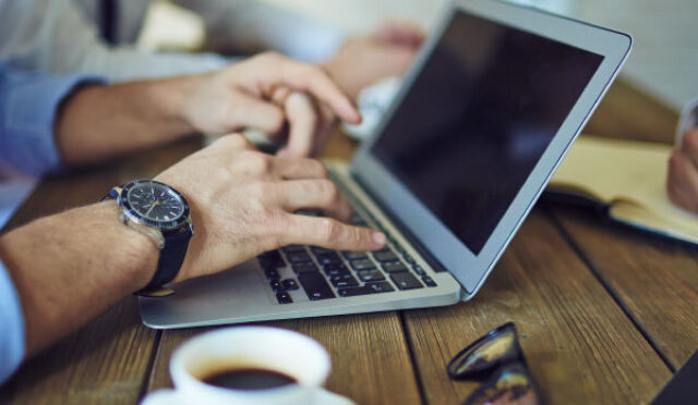 Hands typing on a laptop with a hand nearby writing in a notebook all on a wooden table