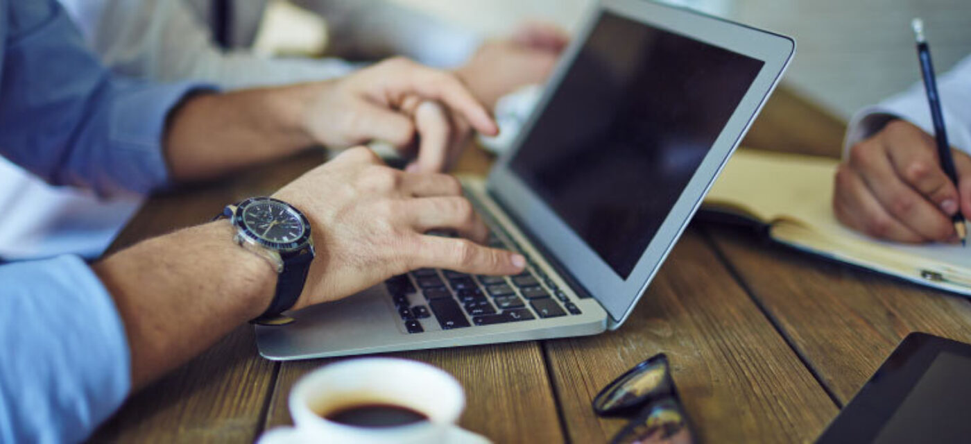 Hands typing on a laptop with a hand nearby writing in a notebook all on a wooden table