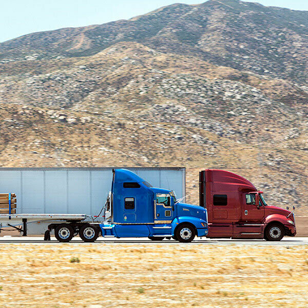 A red semi truck hauling a trailer next to a blue semi truck with hauling a flatbed trailer with cargo on a highway.