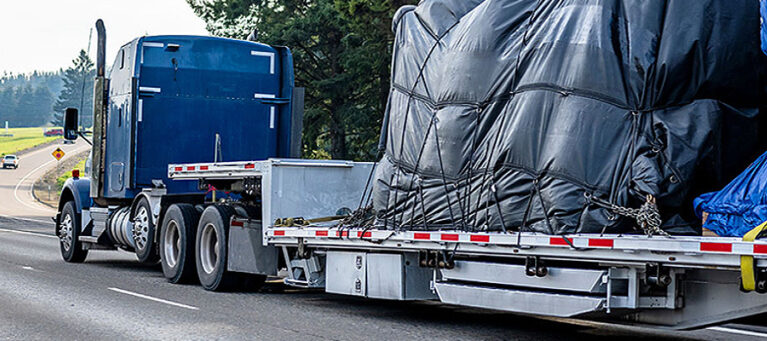 A blue semi-truck hauling a flat bed with a tarp-covered load on a highway.
