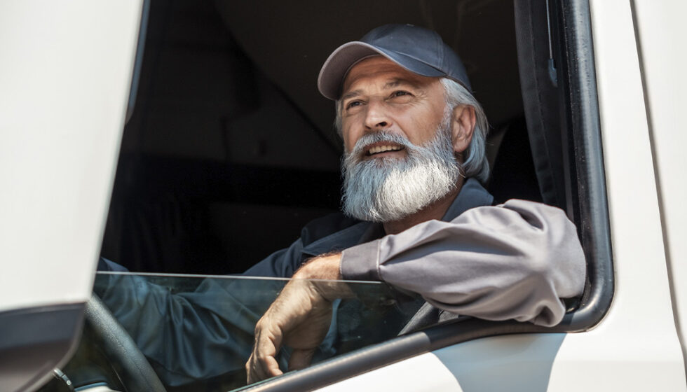 An aged truck driver looking out his driver-side window with his arm resting on the rolled-down window